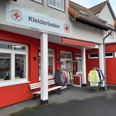 Außenfront des Kleiderladens mit Sitzbank und einigen außenstehenden Regalen mit Bekleidung.