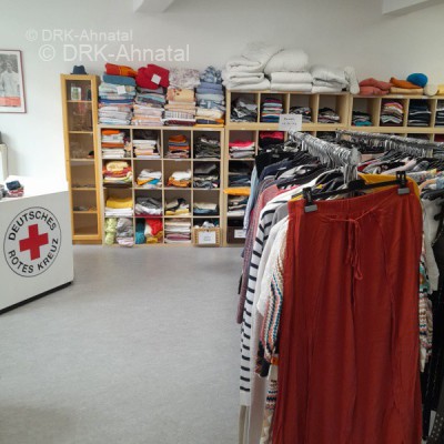 Innenansicht Kleiderladen: An der Wand gegenüber des Eingangs befindet sich ein langes Regal, mit kleineren Textilien und Oberbekleidung. In der Raummitte sieht man ein Regal mit Hosen und Pullovern. Im Linken Bildbereich ist der Tresen abgebildet. 